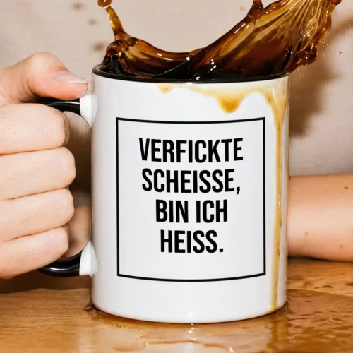 White mug held by a hand with a bold German quote on a white label: 'VERFICKTE SCHEISSE, BIN ICH HEISS.' coffee spills over the rim onto a wooden table, cups edge visible behind.